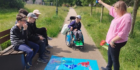 Powiększ grafikę: Chłopcy siedzą na ławce w parku, przed nimi na ziemi leży rozłożona mapa świata. Przed uczniami stoi nauczycielka, ma podniesioną do góry prawą rękę.