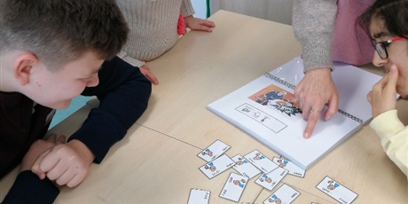 Powiększ grafikę: Nauczyciel pokazuje uczniom miejsce w książce, obok leżą karteczki z symbolami PCS.