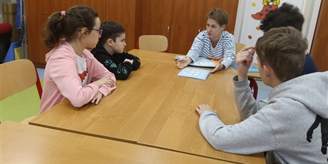 Powiększ grafikę: Uczniowie siedzą przy stole, nauczycielka tłumaczy.