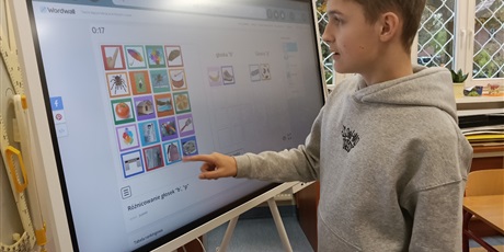 Powiększ grafikę: Chłopiec stoi przy tablicy multimedialnej. Wykonuje zadanie.
