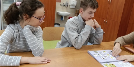 Powiększ grafikę: Chłopiec i dziewczynka siedza przy ławce, patrzą na otwartą stronę skoroszytu.