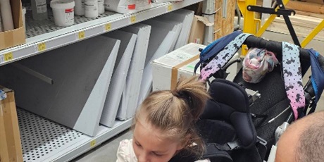 Powiększ grafikę: Dziewczynka na wózku razem z mężczyzną wierci wiertarką w deseczce.