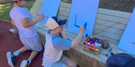 Powiększ grafikę: Uczniowie malują na dużych kartkach opartych o ławki.