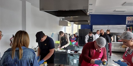 Powiększ grafikę: Widok na salę, uczniowie stoją przy stanowiskach kuchennych, robią kanapki.