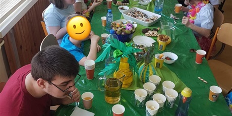 Powiększ grafikę: Uczniowie siedzą przy stole przykrytym zielonym obrusem, na którym stoją kubeczki i ciastka.