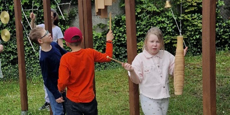 Powiększ grafikę: Dwóch chłopców i dwie dziewczynki grają na instrumentachpowieszonych na drewnianym stelażu na boisku szkolnym.