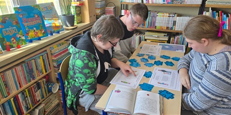 Powiększ grafikę: Dwóch chłopców i dziewczynka siedzą przy stoliku w bibliotece. Na stoliku leżą kawałki niebieskich puzzli i kartki z tekstem.