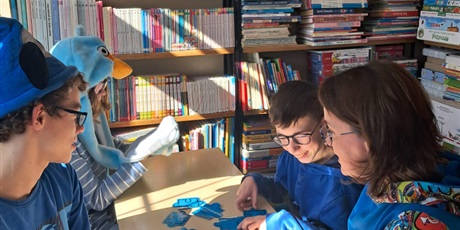 Powiększ grafikę: Dwóch chłopców i dziewczynka ubrani na niebiesko siedzą dookoła stolika w bibliotece między regałąmi z książkami.