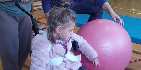 Powiększ grafikę: Dziewczynka na wózku toczy dużą, różową piłkę po ławeczce, obok niej idą dwie panie nauczycielki.