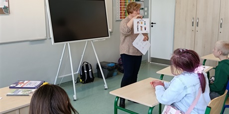 Powiększ grafikę: Kobieta pokazuje uczniom siedzącym przy stolikach planszę.