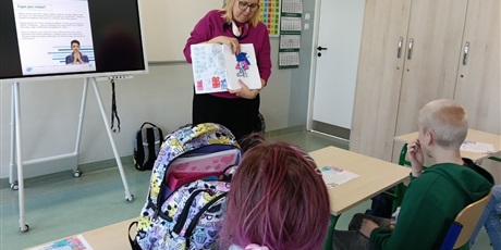 Powiększ grafikę: Kobieta pokazuje uczniom ilustrację w książce.