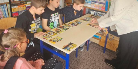Powiększ grafikę: Trzech chłopców i dziewczynka na wózku siedzą przy ławce, na której leżą kawałki duzych puzzli. Obok ławki stoi nauczycielka.