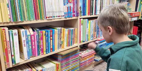 Powiększ grafikę: Chłopiec w zielonej bluzie stoi na tle regałów z książkami.