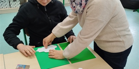 Powiększ grafikę: Uczeń siedzi przy stoliku, nauczyciel stoi obok, pomaga uczniowi przykleic tekst na zieloną kartkę.