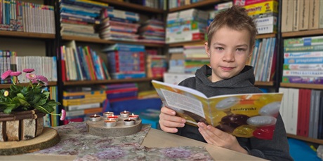 Powiększ grafikę: Chłopiec siedzi przy stoliku w bibliotece, trzyma w rękach otwartą książkę. Na stoliku stoi doniczka ze stokrotkami i świeczki.