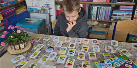 Powiększ grafikę: Chłopiec siedzi przy stoliku w bibliotece, podpiera ręką brodę. na stoliku leżą rozłożone karty i stoi doniczka z stokrotkami.