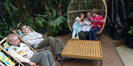 Powiększ grafikę: Trzy dziewczynki siedza na fotelu w kształcie kosza, a dwóch chłopców leży na leżakach w otoczeniu roślin zielonych.