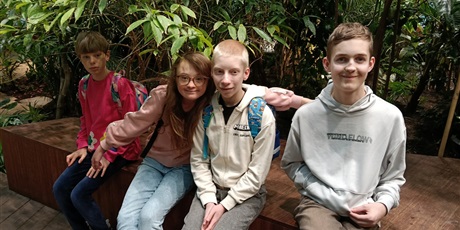 Powiększ grafikę: Czwórka uczniów siedzi na podeście pod zielonymi krzewami.