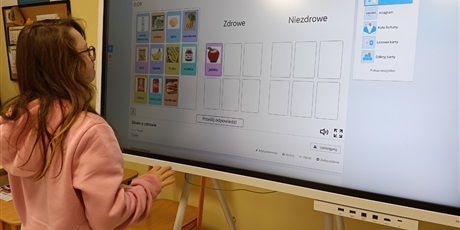 Powiększ grafikę: Dziewczynka stoi przed tablicą multimedialną z zadaniem.