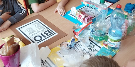 Powiększ grafikę: Na stole leżą kartkiw kolorach odpowiadających pojemnikom na śmieci, na kartkach stoją plastikowe butelki, leżą kawałki papieru, zakrętki.
