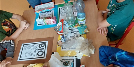Powiększ grafikę: Uczniowie siedzą dookoła stołu, na którym na kartkach w kolorach pojemników do śmieci leżą różne rodzaje śmieci. 