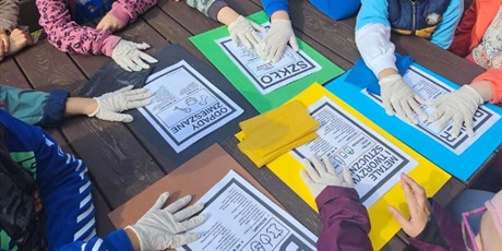 Powiększ grafikę: Uczniowie siedzą przy stole piknikowym w ogrodzie, na stole leżą kartki w kolorach pojemników na śmieci.