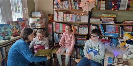 Powiększ grafikę: Troje uczniów siedzi w półkole w bibliotece i ogląda prezentowaną ptrzez nauczyciela książkę.