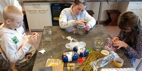 Powiększ grafikę: Dzieci siedzą przy stole, na którym leżą rożne elementy dekoracykne. Ozdabiają słoiki.