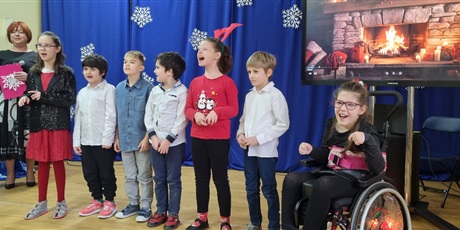 Powiększ grafikę: Uczniowie stoją natle świątecznej dekoracji i śpiewają.