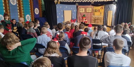 Powiększ grafikę: Uczniowie siedzą w sali gimnastycznej, widać scenę, na scenie aktorka w rudej peruce i zielonym kapeluszu.
