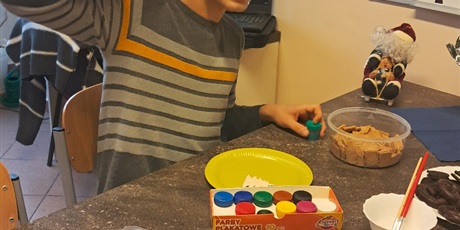 Powiększ grafikę: Chłopiec siedzi przy stole, na którym leżą pierniki i farby.