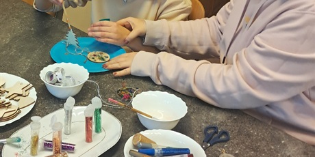 Powiększ grafikę: Uczennica wraz z nauczycielką maluje choinkę wyciętą z drewna, na stole leżą różne przybory plastyczne.