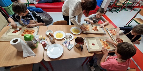 Powiększ grafikę: Uczniowie siedzą przy stole i smarują spody pizzy sosem pomidorowym.