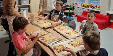 Powiększ grafikę: Chłopcy siedzą dookoła stołu przed nimi przygotowane do pieczenia spody pizzy na blachach.