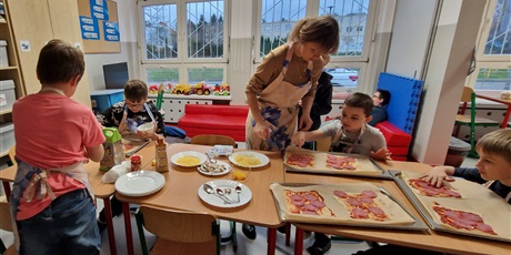 Powiększ grafikę: Uczniowie siedzą przy stole, układaja na spodach pizzy wędlinę, obok stoi nauczyciel.