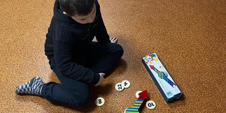 Powiększ grafikę: Chłopiec siedzi na podłodze, przed nim leżą cyferki na kartonikach i strzałka.