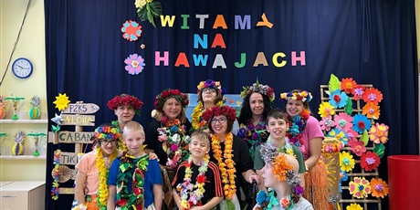 Powiększ grafikę: Grupa uczniów z wiankami na głowach i kwiatowymi koralami stoi na granatowym tle z napisem "Witamy na Hawajach".