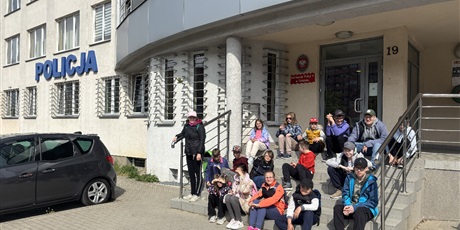 Powiększ grafikę: Grup uczniów siedzi na schodach przed budynkiem komisariatu policji.
