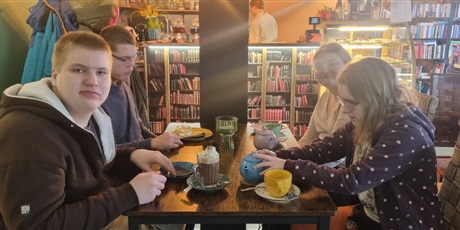 Powiększ grafikę: Czworo uczniów siedzi przy stoliku w kawiarni, piją herbatę, jedzą ciastko, w tle regały z książkami.