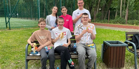 Powiększ grafikę: Trzech uczniów siedzi na ławce na boisku, dwie dziewczynki i chłopiec stoją za nimi.