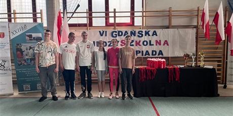 Powiększ grafikę: Uczniowie stoją w rzędzie na sali gimnastycznej na tle baneru z nazwą zawodów, obok nich stoi stolik z medalami i pucharami.