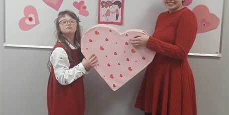 Powiększ grafikę: Dwie dziewczynki stoją na tle napisu "Walentynki". Trzymają razem duyży, różowy karton w kształcie serca.