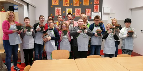 Powiększ grafikę: Grupa uczniów z nauczycielami stoi na tle tablicy z jesiennymi dekoracjami, wszyscy trzymaja w rękach doniczki z wrzosami.