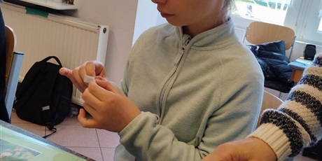 Powiększ grafikę: Dziewczynka siedzi przy stole, na którym leżą zielone kartki i piktogramy, odkleja folię od karteczki.