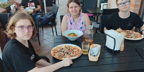 Powiększ grafikę: Dwie dziewczynki i chłopiec siedzą przy stoliku w restauracji. Dziewczynka i chłopiec mają przed sobą pizze, druga dziewczynka danie w głębokim talerzu.