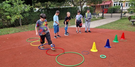 Powiększ grafikę: Uczniowie stoją w rzędzie, rzucają ringo na pachołki ustawione przed nimi. jeden uczeń chodzi między rozłożonymi kołami hulahop.