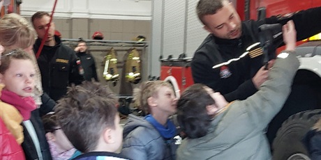 Powiększ grafikę: Uczniowie stoją obok wozu strażackiego, mężczyzna w stroju strażaka pokazuje im wyposażenie wozu.