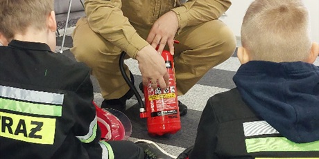 Powiększ grafikę: Mężczyzna w kombinezonie strażaka pokazuje gaśnicę uczniom siedzącym przed nim.