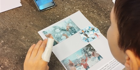 Powiększ grafikę: Chłopiec siedzi przy stole, na którym leży kartka z nalepionymi zimowymi obrazkami, patrzy na zdjęcie w telefonie.