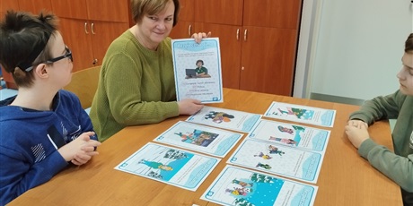 Powiększ grafikę: Dwóch chłopców siedzi naprzeciwko siebie przy stole, obok jednego z nich kobieta trzyma podniesioną kartkę z rysunkiem i tekstem. Na stole leżą inne kartki.